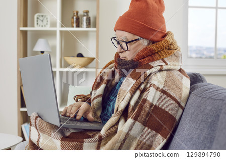 Elderly man working at home with laptop and trying to warm up with heating problems. 129894790