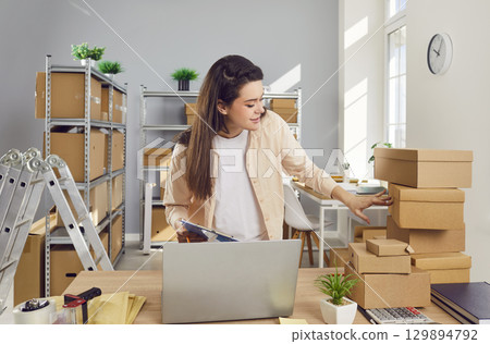 Young confident female employee working in warehouse among cardboard boxes. Young confident female employee working in warehouse among cardboard boxes. 129894792