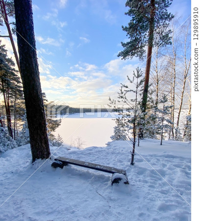 Wooden bench overlooking frozen lake in winter forest Wooden bench overlooking frozen lake in winter forest 129895910