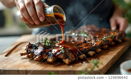 A person pouring barbecue sauce on grilled food at a summer barbecue, set against a lively outdoor backdrop 129896642