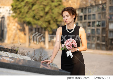 Sad woman in black dress with bouquet of flowers and handkerchief in hands, stands in cemetery 129897285