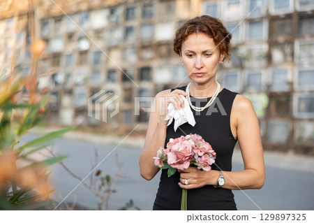 Sorrowful woman in black standing in cemetery with flowers 129897325