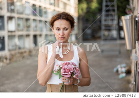 Sad woman in white croptop with bouquet of flowers and handkerchief in hands, stands in cemetery 129897418