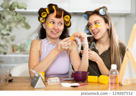 Two besties in colorful hair rollers applying under-eye patches 129897552