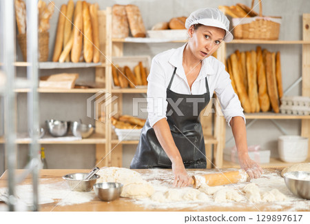 Making dough in bakery - female baker rolling out dough with rolling pin Making dough in bakery - female baker rolling out dough with rolling pin 129897617