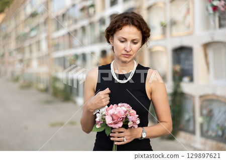 Drieving woman in black dress standing with bouquet by columbarium 129897621