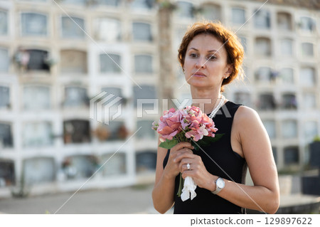 Sad woman in black dress with bouquet of flowers and handkerchief in hands, stands in cemetery 129897622