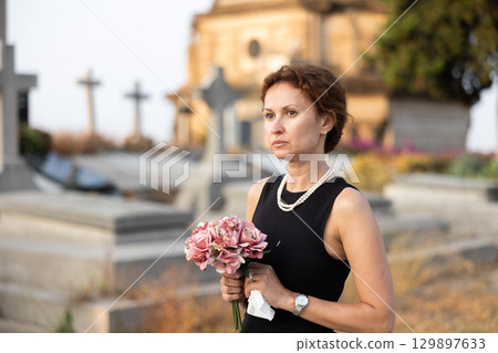 Sad woman in black dress with bouquet of flowers and handkerchief in hands, stands in cemetery 129897633