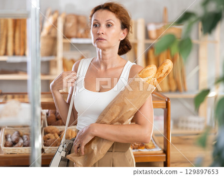 Woman with baguettes in paper bag stands near glass showcase in bakery 129897653