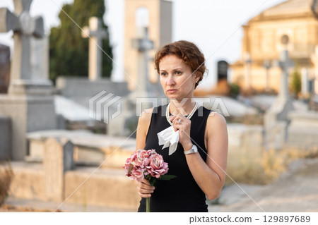 Grieving woman in black standing in cemetery with flowers 129897689