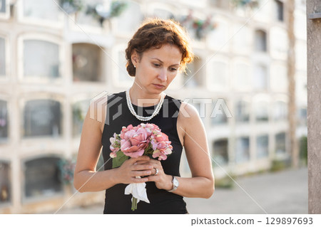 Sad woman in black dress with bouquet of flowers and handkerchief in hands, stands in cemetery 129897693