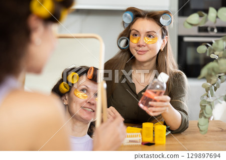 Woman in hair rollers applying eye patches in front of mirror with friend 129897694
