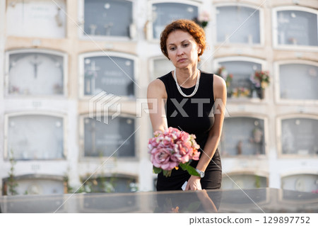 Sorrowful woman puts bouquet of hydrangeas on grave of deceased friend, mourns loss 129897752