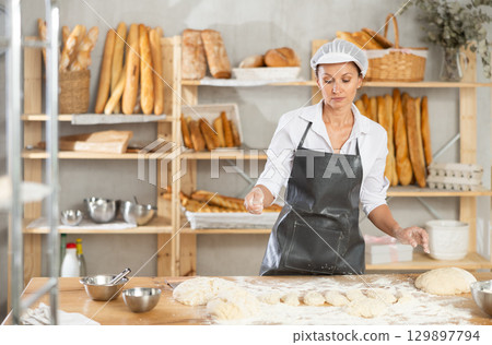 Woman works in bakery as baker, cuts dough into portions, forms pieces of dough to create buns. Woman works in bakery as baker, cuts dough into portions, forms pieces of dough to create buns. 129897794