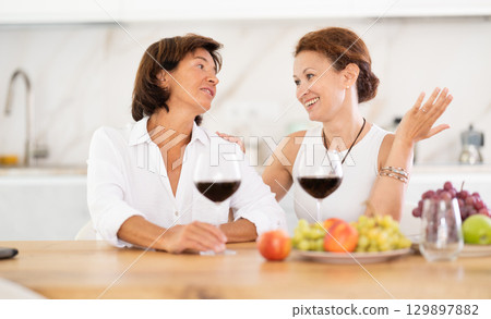 Two girlfriends chatting and drinking red wine in cozy home kitchen Two girlfriends chatting and drinking red wine in cozy home kitchen 129897882