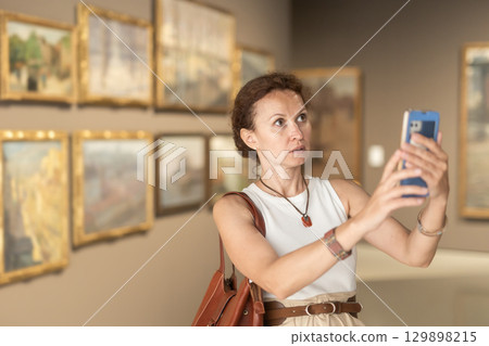 Interested woman looking closely at paintings in art gallery, using smartphone and taking photo 129898215