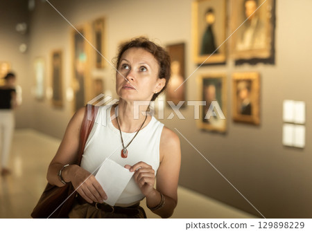 Thoughtful attentive woman standing in front of painting in art museum 129898229