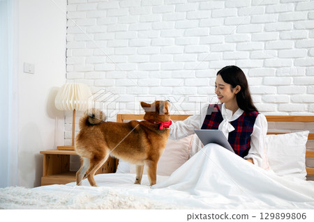 Smiling woman sitting in bed with a tablet, happily petting her dog wearing a red bow in a cozy bedroom Smiling woman sitting in bed with a tablet, happily petting her dog wearing a red bow in a cozy bedroom 129899806