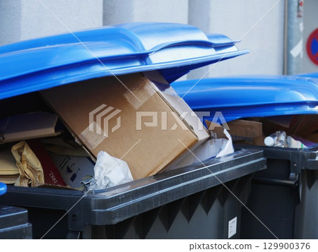 Blue trash can is full of trash, including a box and a roll of toilet paper 129900376