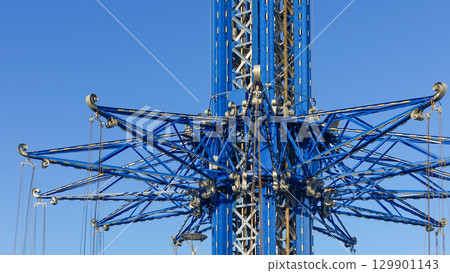 Towering amusement park ride structure with intricate blue metal framework against clear blue sky, showcasing engineering and design excellence 129901143
