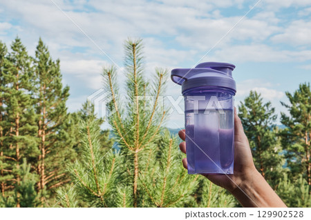 Female hand holding bottle of water against coniferous trees and blue sky Female hand holding bottle of water against coniferous trees and blue sky 129902528