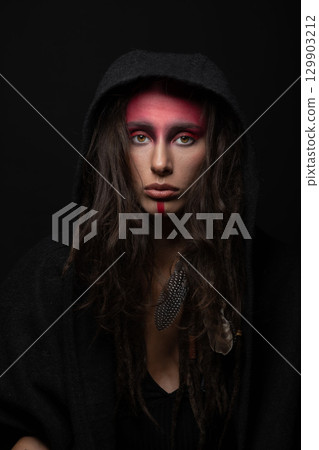 Model with tribe red make-up, dreadlocks, feather attached to her hair hiding under hood Model with tribe red make-up, dreadlocks, feather attached to her hair hiding under hood 129903212