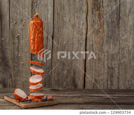 Floating slices of smoked sausage with falling peppercorns on rustic wooden background. copy space. 129903734