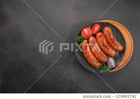 Grilled sausages with tomatoes, onion, and parsley served on black plate with wooden board. top view, copy space Grilled sausages with tomatoes, onion, and parsley served on black plate with wooden board. top view, copy space 129903742