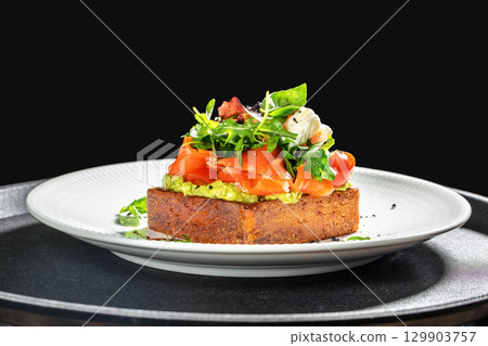 Gourmet open sandwich with smoked salmon, avocado spread, cherry tomatoes, fresh greens, and soft cheese served on a thick slice of rustic bread on a white plate 129903757
