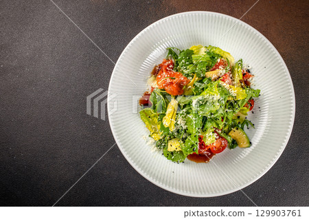 Fresh green salad with arugula, lettuce, avocado, cherry tomatoes, and grated cheese, served with dressing on a white plate over a dark background, top view 129903761