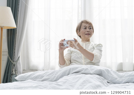 Senior woman operating a smartphone on her bed in her bedroom 129904731