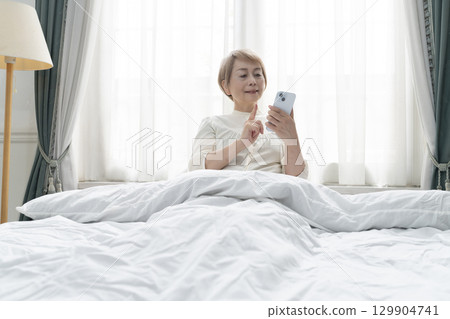 Senior woman operating a smartphone on her bed in her bedroom 129904741