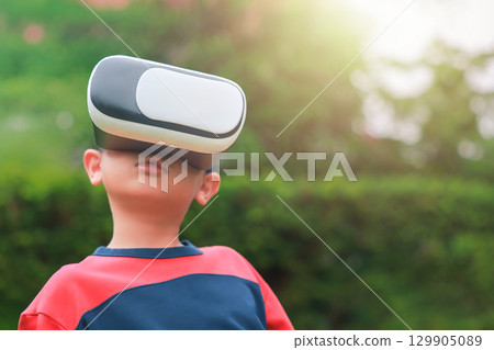 Young boy wearing virtual reality headset outdoors, child experiencing VR technology in nature, kid playing immersive digital game with modern gadget. Virtual reality while sitting outdoors. Young boy wearing virtual reality headset outdoors, child experiencing VR technology in nature, kid playing immersive digital game with modern gadget. Virtual reality while sitting outdoors. 129905089