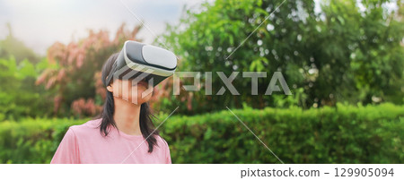 Young woman wearing virtual reality headset outdoors, female exploring VR technology in nature, person experiencing immersive digital world with modern gadget and innovation. 129905094