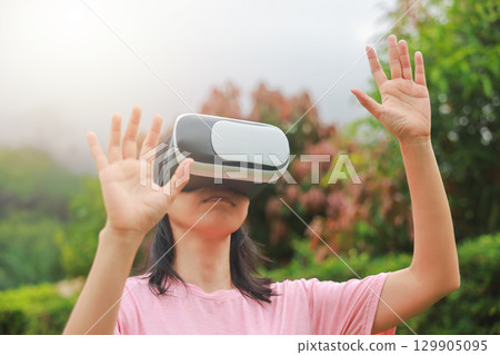Young woman wearing virtual reality headset outdoors, female exploring VR technology in nature, person experiencing immersive digital world with modern gadget and innovation. 129905095