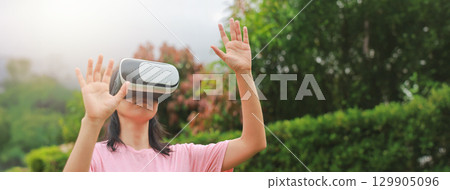Young woman wearing virtual reality headset outdoors, female exploring VR technology in nature, person experiencing immersive digital world with modern gadget and innovation. 129905096
