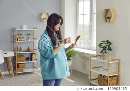 Young pregnant woman standing at home and holding a comb with her hair falling out. 129905485