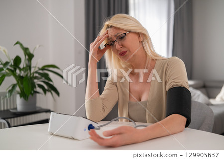 Blonde middle aged woman in beige clothes holding blood pressure monitor, appearing uncomfortable and worried due to headache in modern living room Blonde middle aged woman in beige clothes holding blood pressure monitor, appearing uncomfortable and worried due to headache in modern living room 129905637