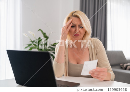 Portrait of blonde middle aged woman sitting in cozy living room, sadly checking bill and struggling with household budget during financial crisis 129905638