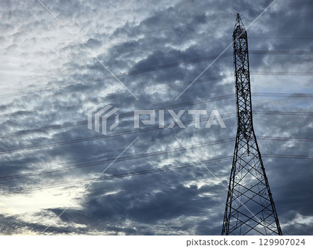 A towering electricity pylon stands silhouetted against a dramatic, cloudy sky, 129907024