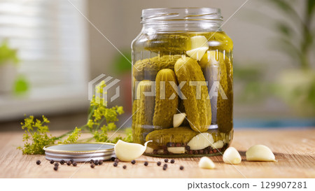 Glass jar filled with pickled cucumbers, garlic and spices on wooden table in natural kitchen light, homemade food preservation concept 129907281