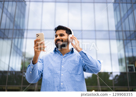 A smiling man wearing headphones and holding a smartphone in front of a modern glass building. 129908355
