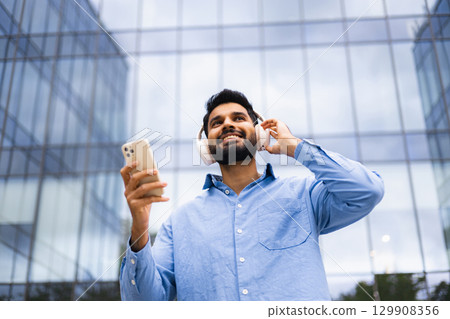A smiling man listens to music on his phone while wearing headphones, standing in front of a modern glass building. 129908356