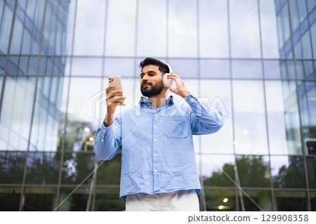 A man in a blue shirt and white pants stands outdoors, holding a phone and wearing headphones in front of a modern building. 129908358