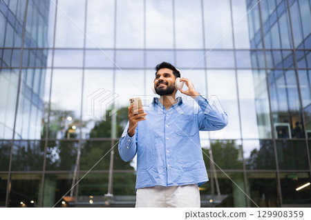 A man in a blue shirt listens to music on headphones while holding a phone, standing outside a modern building. 129908359