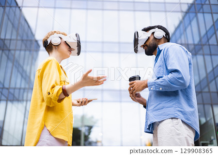 Two people wearing VR headsets interact outside a modern glass building, exploring virtual reality with coffee and a phone. Two people wearing VR headsets interact outside a modern glass building, exploring virtual reality with coffee and a phone. 129908365