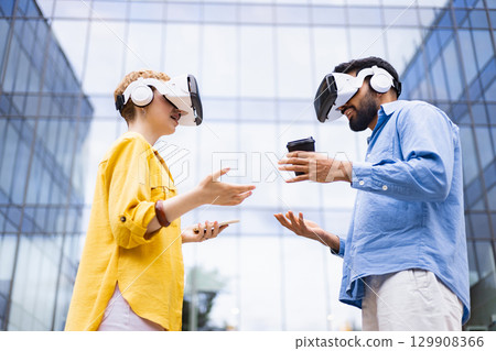 Two people wearing VR headsets interact outdoors, one holding a coffee cup and the other a phone, set against a modern building. 129908366