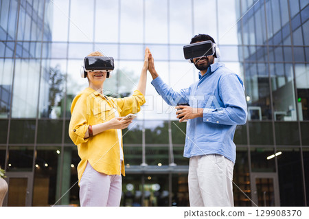 Two people wearing VR headsets high-fiving, standing in front of a modern building, symbolizing technology and teamwork. 129908370