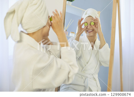 Woman in bathrobe making face mask putting slices of cucumbers on eyes and looking at mirror Woman in bathrobe making face mask putting slices of cucumbers on eyes and looking at mirror 129908674