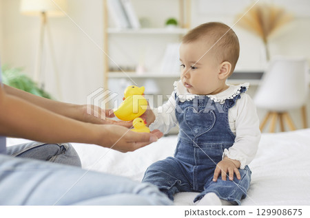 Curious baby in denim overalls reaching for yellow rubber duck toys during playtime with mother. Curious baby in denim overalls reaching for yellow rubber duck toys during playtime with mother. 129908675